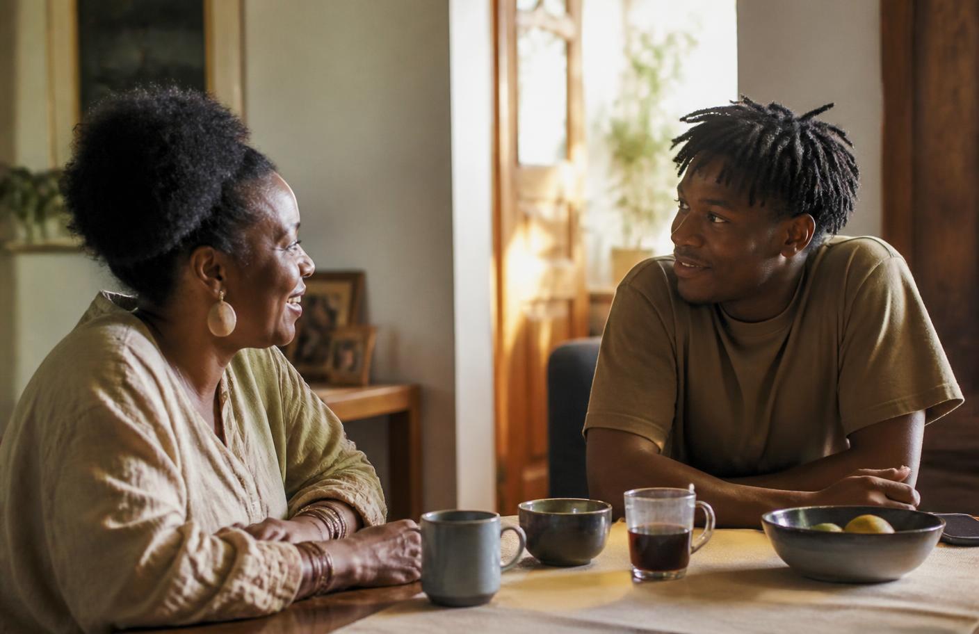An older woman and a younger man sitting across a table in a sunlit room, talking.